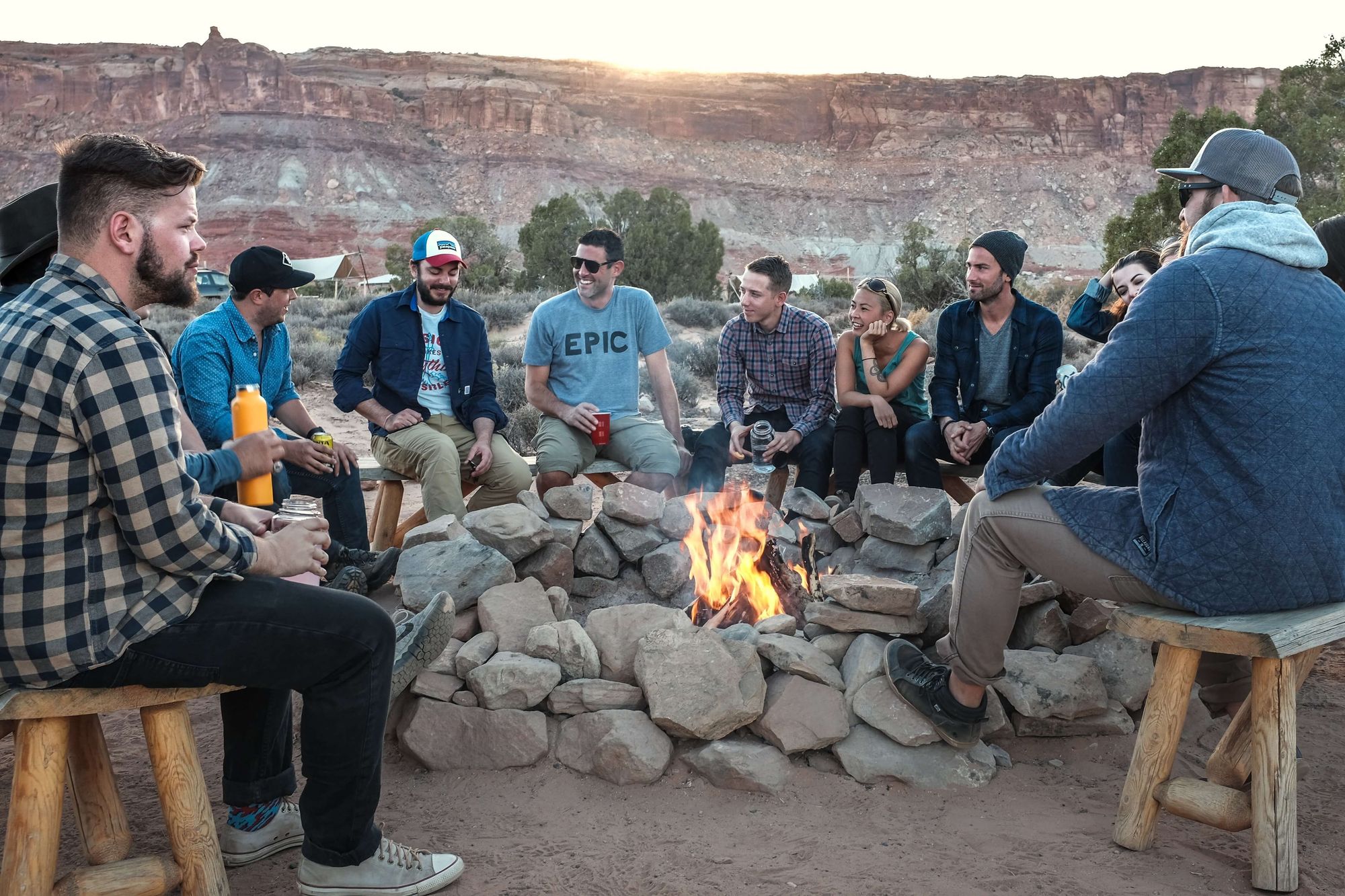 A group of campers laughing & socializing around a fire, in the daytime, with a canyon in the background.