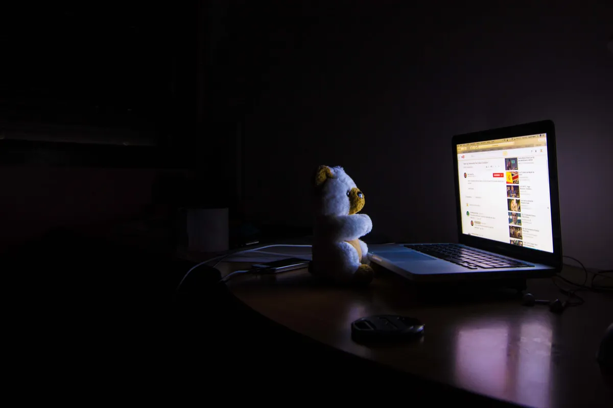 A toy teddy bear in front of a bright laptop on a desk at night.