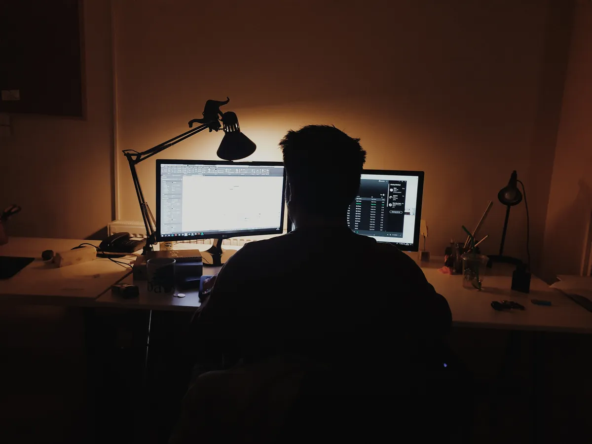 A man working at a PC desktop workstation with two monitors in a dimly lit room. 