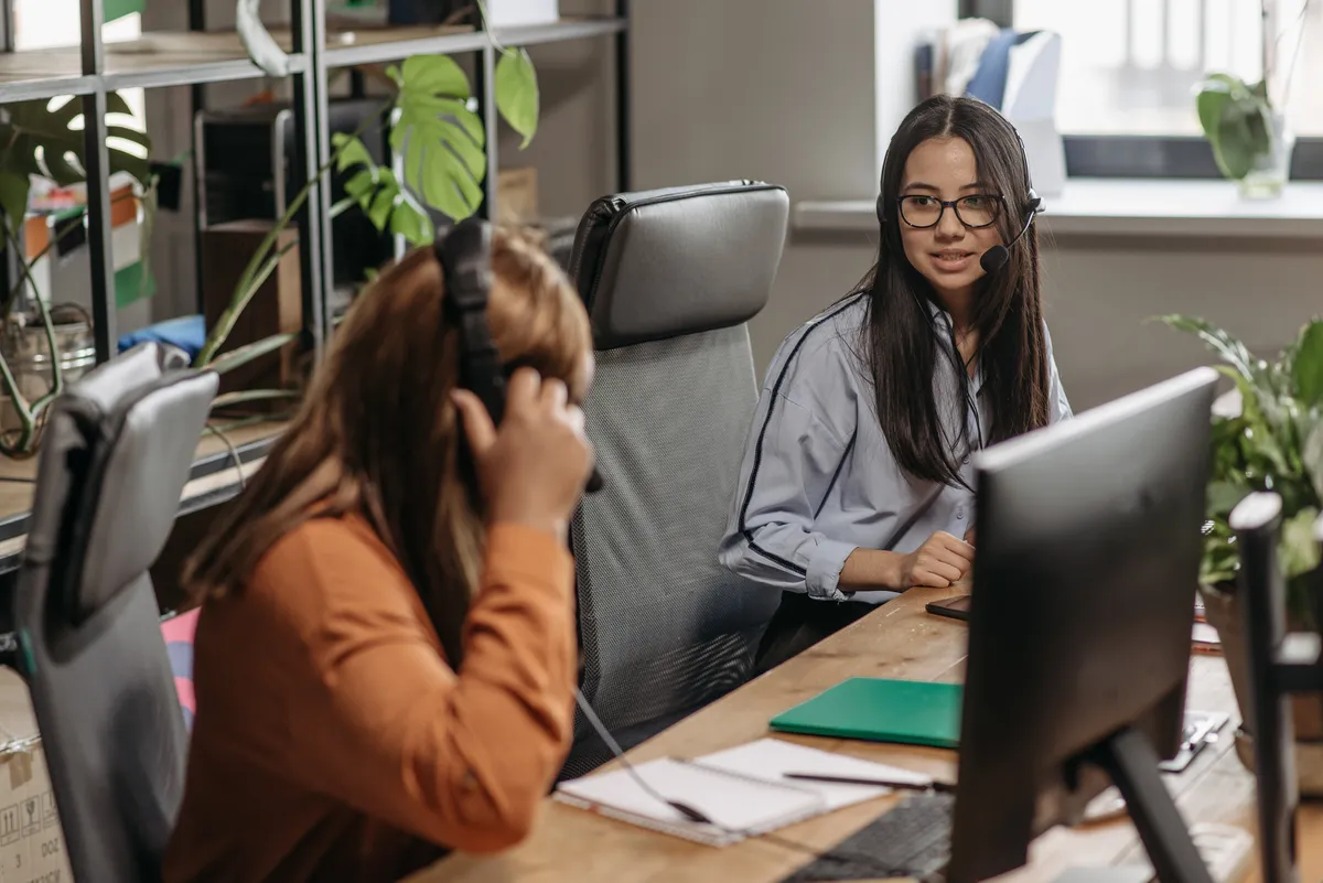 Two women wearing headphones working at desks with computers in an open workspace.